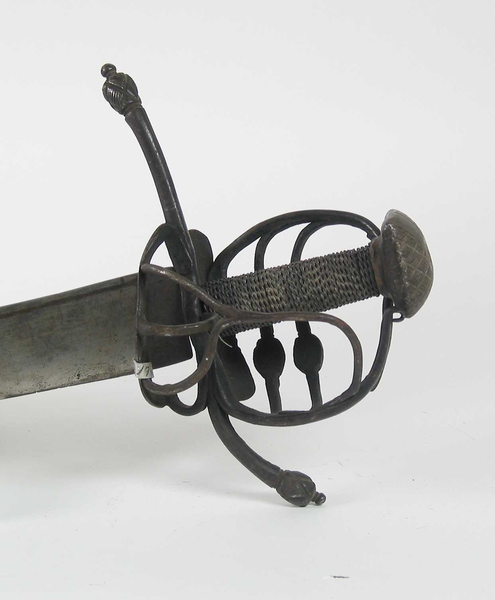 Pyramideformet knapp med strekdekor. Asymmetrisk kurvfeste med to sidebøyler( øverste avbrukket )med riflede fortykkelser på midten. mellom håndbøylen og kurvens ytterkant. Innsiden har en  sidebøyle. Kurvens basis er på utsiden en kvadratisk parérplate med to hjerteformede gjennombrytninger. På innsiden er det en klingebrekker og tommelbøyle. Opp og nedbøyde parérstenger med riflede fortykkelser på endene.
Tregrep med nyere jerntrådvikling.
Enegget krum klinge med 3 hull ipå klingspissen og en smal hulslipning på ryggsiden.Tre smedmerker på venstre side. 