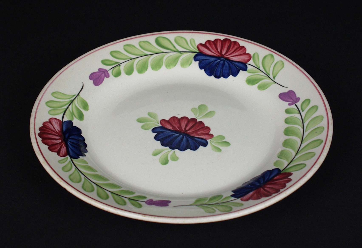 Deler av servise i steintøy og flintgods. Godset er hvitt med malt blomsterdekor. Rød og blå blomst i midten med en ranke av grønne blad.