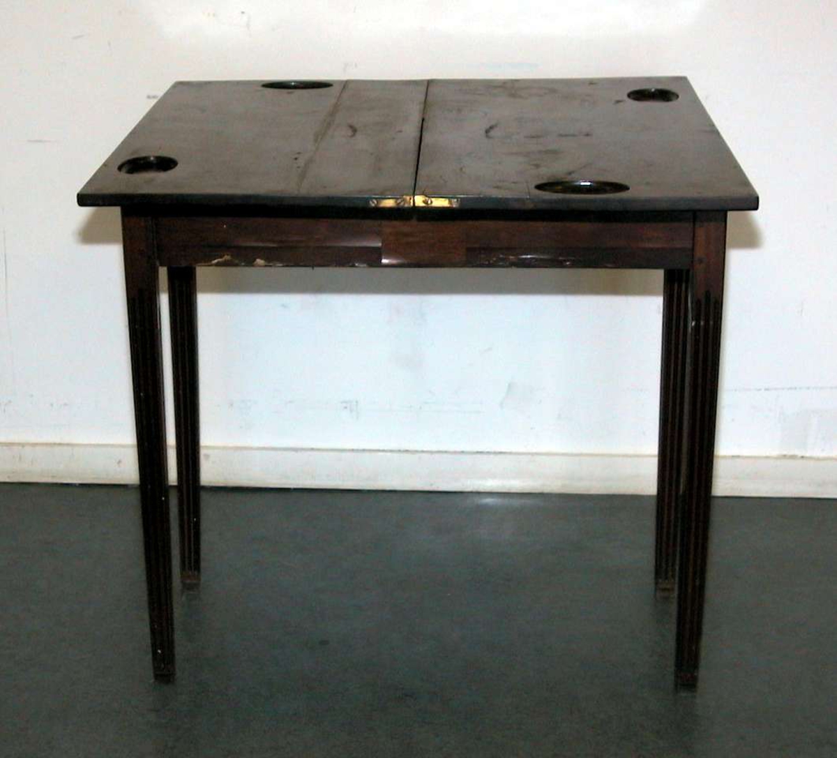 Spillebord med utslagbar plate. Det er et rom under bordplaten. Platen har fire fordypninger. Bordet har rette ben med rifler. Fineringen på en side av sargen er skadet. Bordet er brunpolert.