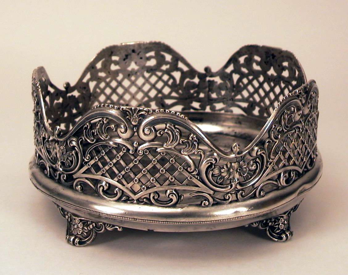 Fat i metall, antagelig tinn. Fatet står på fire ben og har korpus med  gjennombrutt, støpt gitter og blomsterdekor. Fatet mangler innmat i glass. Det er hull i bunnen av fatet.