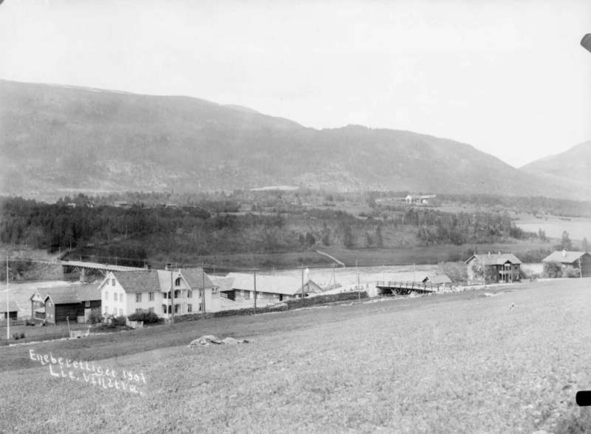 12.06.1907. N. Byhre, Sundbroen og Præstgaarden. Boliger, uthus, elv ,bro, åsside med trær, dyrket mark.