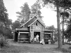 Makarna Hydén med döttrar och gäster vid sommarbostaden Talludden i Sandvik. Upplysningsvis uppfördes huset ursprungligen på den så kallade Kreatursvallen i Linköping i samband med 1898 års lantbruksmöte i staden, men flyttades därefter till denna natursköna plats med utsikt över sjön Roxen. På trappan ser vi järnhandlare Mauritz Hydén med makan Maria Sofia och döttrarna Anna samt Lisa. Vid bordet sitter kamrer Axel Lundblad, dennes dotter Axeline och Gottfrid Söderberg, den sistnämnda svåger respektive bror till makarna Hydén. Fotoår omkring förra sekelskiftet.