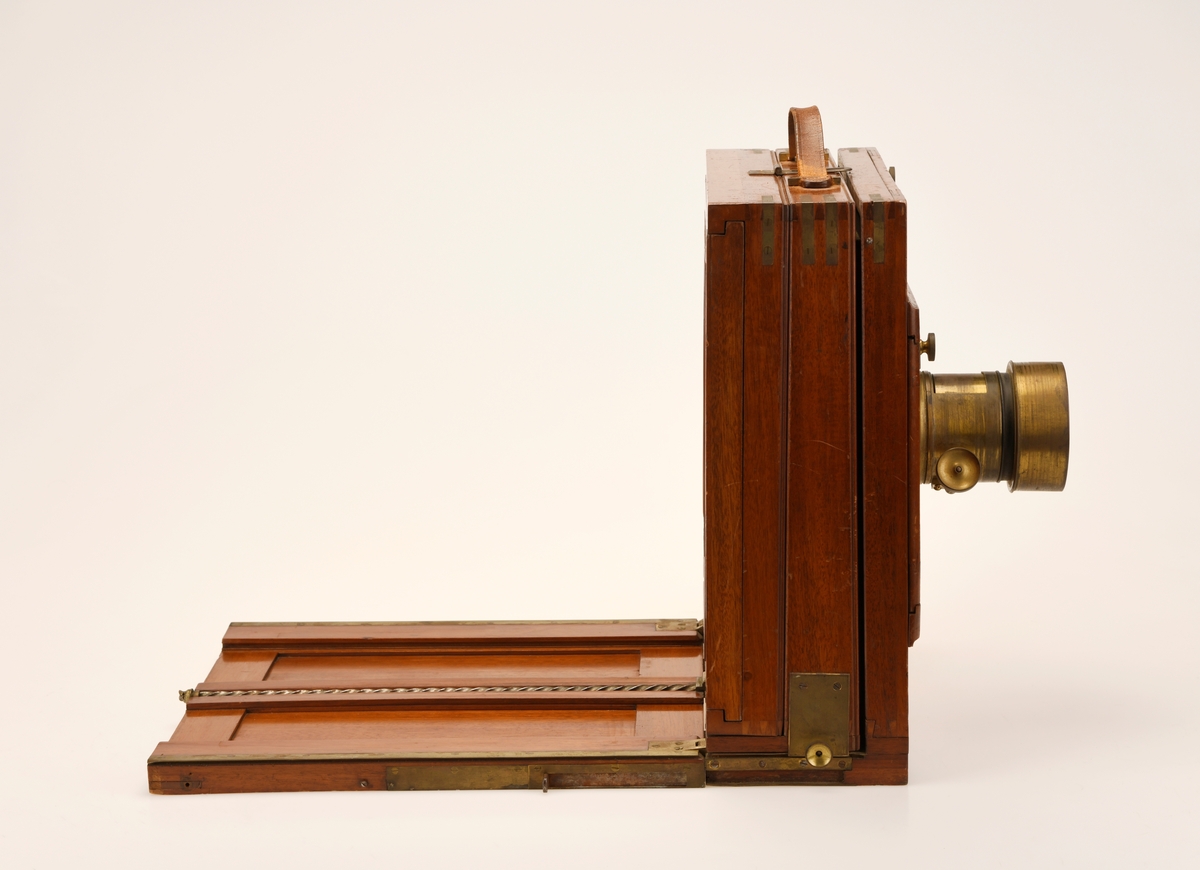 Et belgkamera i lakkert tre. Kamerahuset kan flyttes fram og tilbake langs en skinne mens fronten med objektivplaten står i ro. Platen med skinner på kan brettes opp for å beskytte mattskiven og gjøre kameraet mer kompakt. Objektivplaten kan beveges i høyden og til siden. Objektivet kan dras ut med et hjul på objektivet. Det har beslag i messing. Mattskiven er på 25x25 cm og kan skyves til siden for å sette inn en filmplate. Håndtak i skinn på toppen.