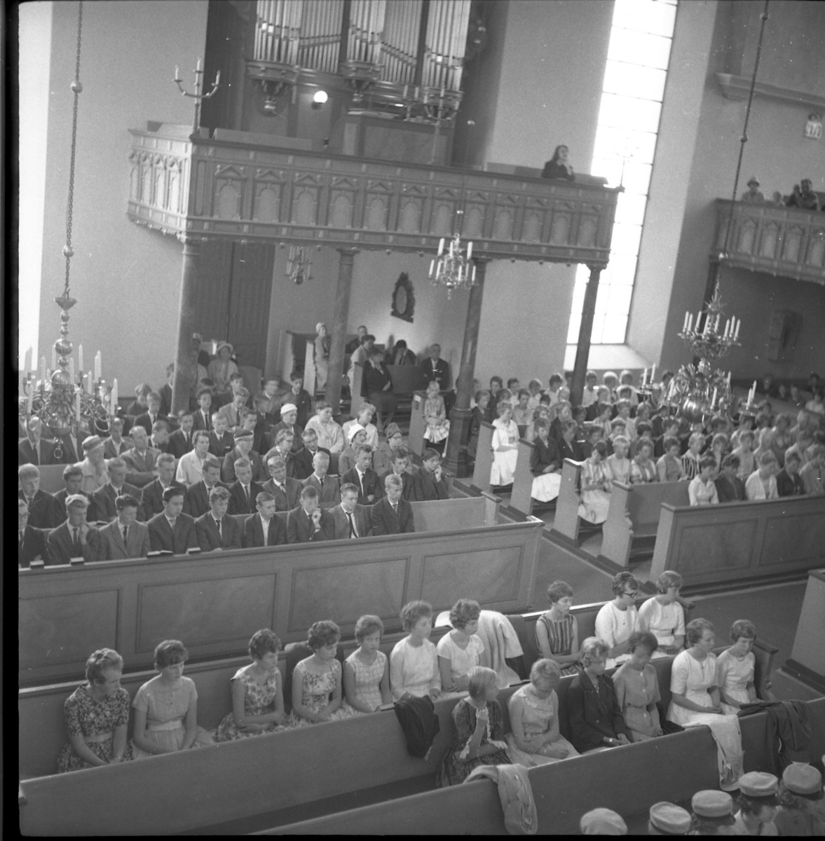 Bänkradera i Gränna kyrka är fullsatta med unga och gamla personer. Man kan skymta att personerna på de främre raderna bär realskolemössa. Flickorna är klädda i mönstrade klänningar med snäv midja och vid kjol. De flesta har korta, lockiga frisyrer. Killarna har kavaj och slips. På läktaren över entrédörren står orgeln.
