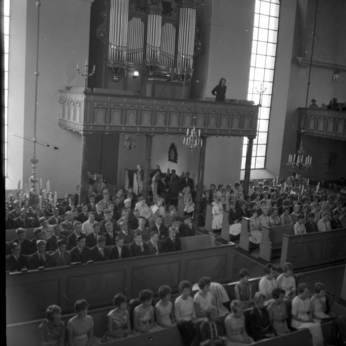 Bänkradera i Gränna kyrka är fullsatta med unga och gamla personer.  Flickorna är klädda i mönstrade klänningar med snäv midja och vid kjol. De flesta har korta, lockiga frisyrer. Killarna har kavaj och slips. På läktaren över entrédörren står orgeln.