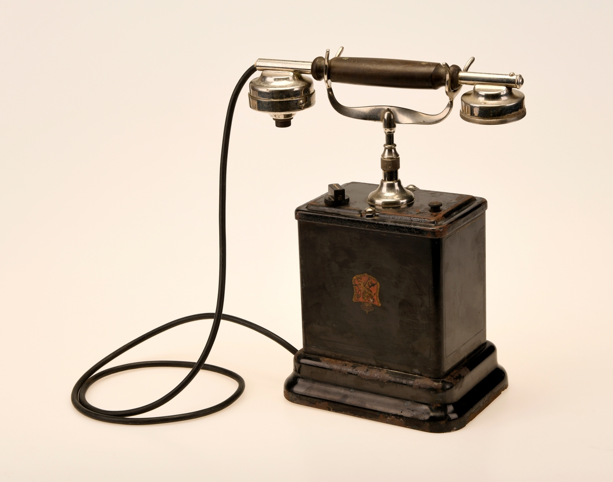 En svart bordtelefon av Elektrisk Bureau sin 1922 modell. "Den norske løve" pryder forsiden og baksiden av aparatkassen, disse er slitt og i tillegg oppned. Forøvrig har telefonen en y-gaffel. Ledningen som går fra aparatkassen til telefonrøret er av rund plast, noe som tyder på at den har blitt byttet ut på et tidspunkt. Sveiven mangler, deler av telefonrøret mangler, og ledningen med støpsel er fjernet.