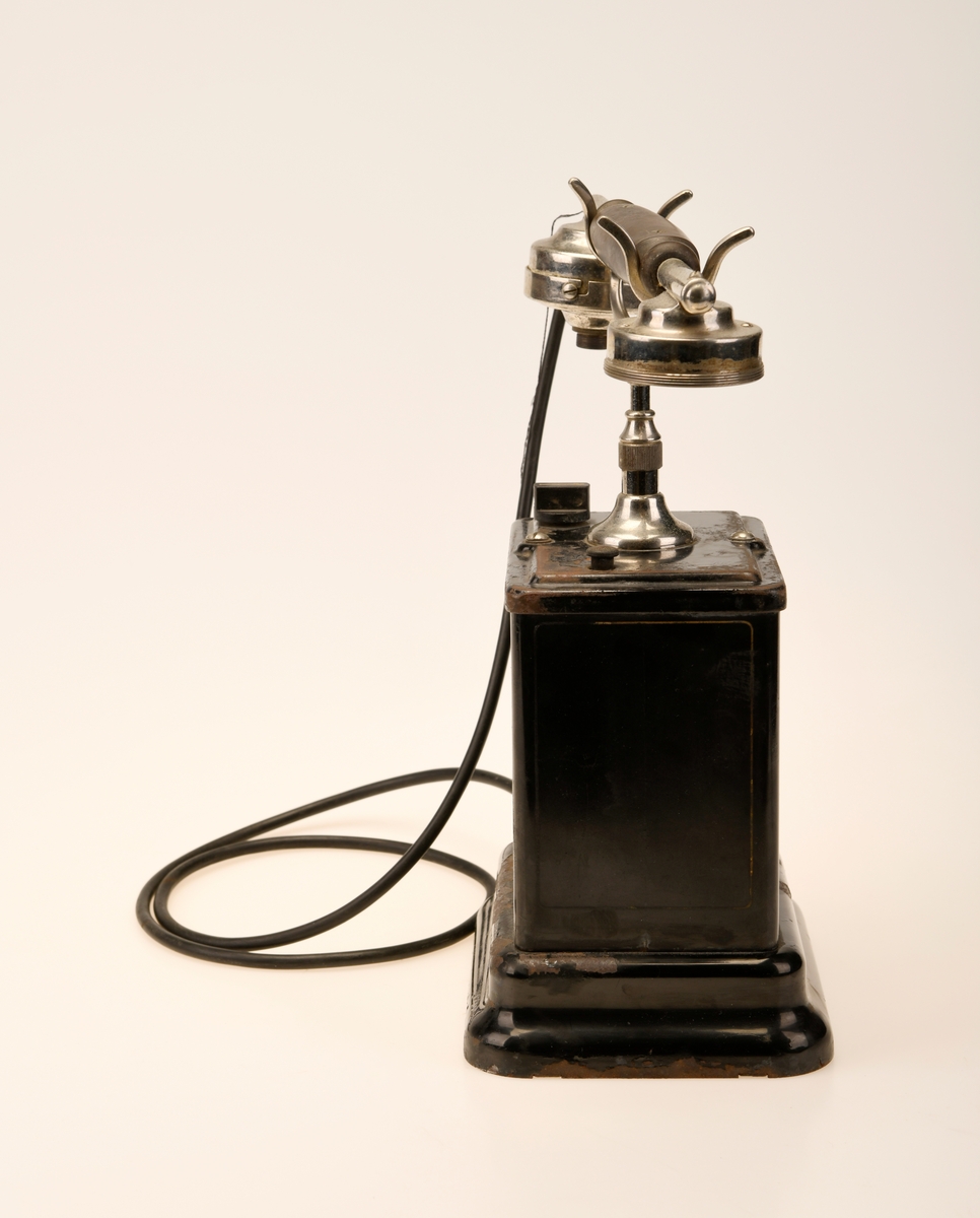 En svart bordtelefon av Elektrisk Bureau sin 1922 modell. "Den norske løve" pryder forsiden og baksiden av aparatkassen, disse er slitt og i tillegg oppned. Forøvrig har telefonen en y-gaffel. Ledningen som går fra aparatkassen til telefonrøret er av rund plast, noe som tyder på at den har blitt byttet ut på et tidspunkt. Sveiven mangler, deler av telefonrøret mangler, og ledningen med støpsel er fjernet.