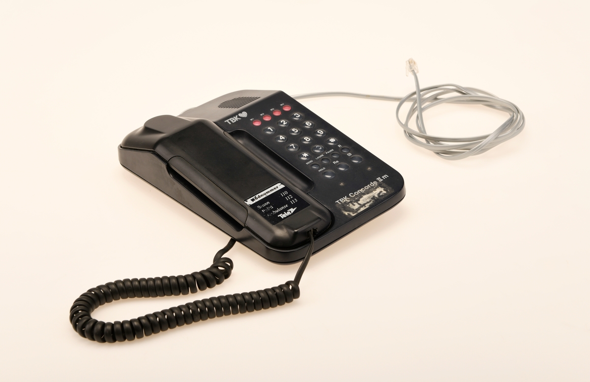 Sort bordtelefon med 23 taster fra TBK. Telefonen er av modelltypen TBK Concorde II m. Telefonen kommer med en svart spirallisert ledning til telefonrøret og en hurtigkopling i grått.
