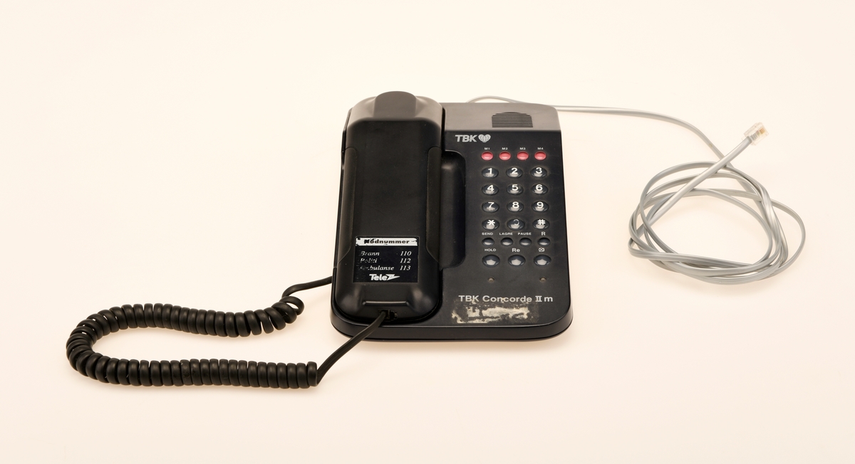 Sort bordtelefon med 23 taster fra TBK. Telefonen er av modelltypen TBK Concorde II m. Telefonen kommer med en svart spirallisert ledning til telefonrøret og en hurtigkopling i grått.