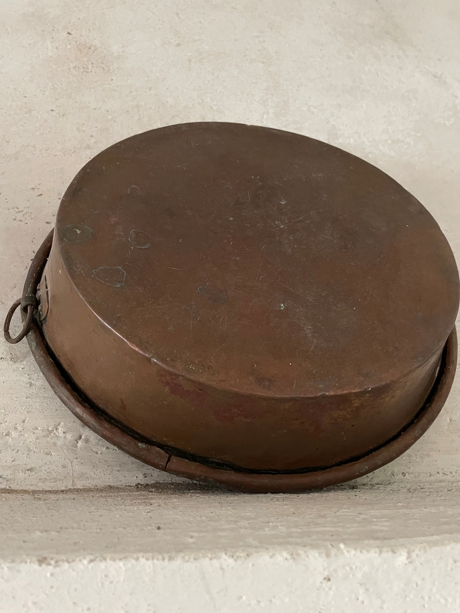 En cirkelformad metallplatta med en rak kant runt om som avslutas med en böjd kant utåt. Under kanten är en ögla fastsatt.