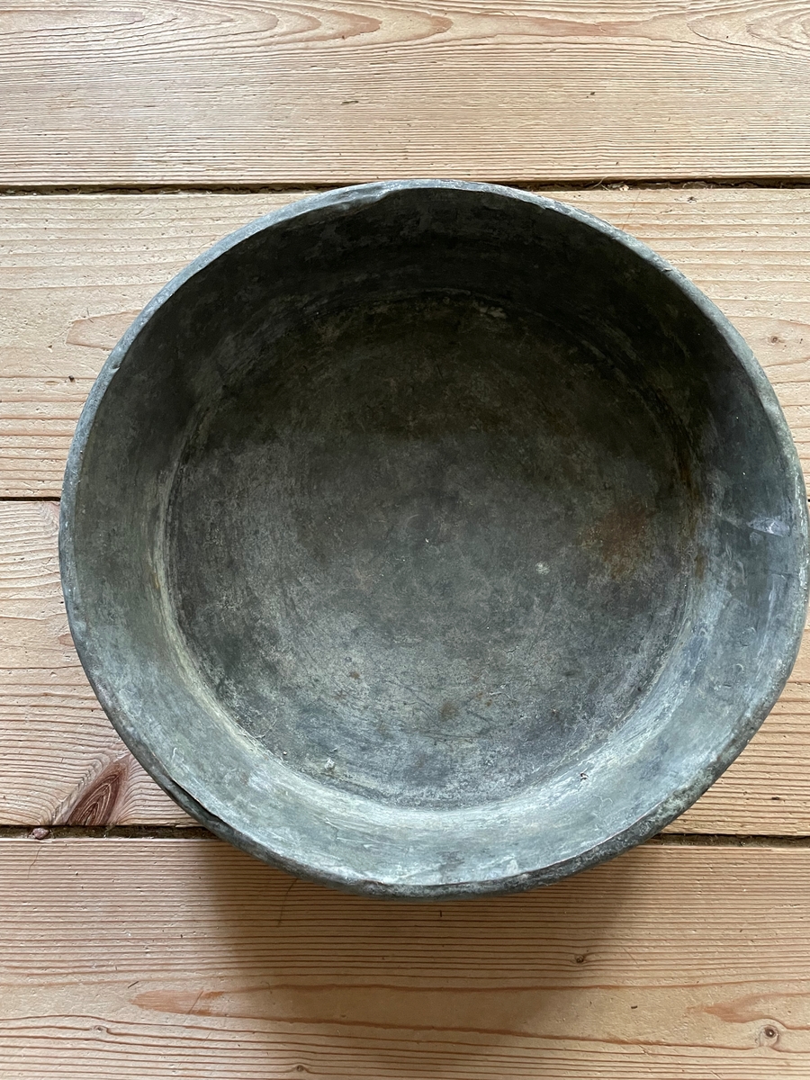 En cirkelformad metallplatta med en rak kant runt om som avslutas med en böjd kant utåt. Under kanten är en ögla fastsatt.