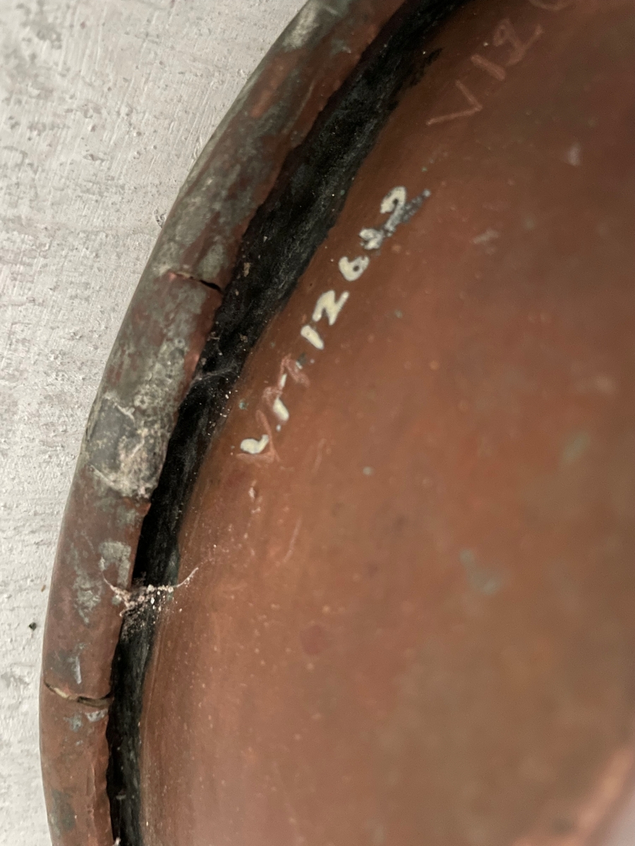 En cirkelformad metallplatta med en rak kant runt om som avslutas med en böjd kant utåt. Under kanten är en ögla fastsatt.