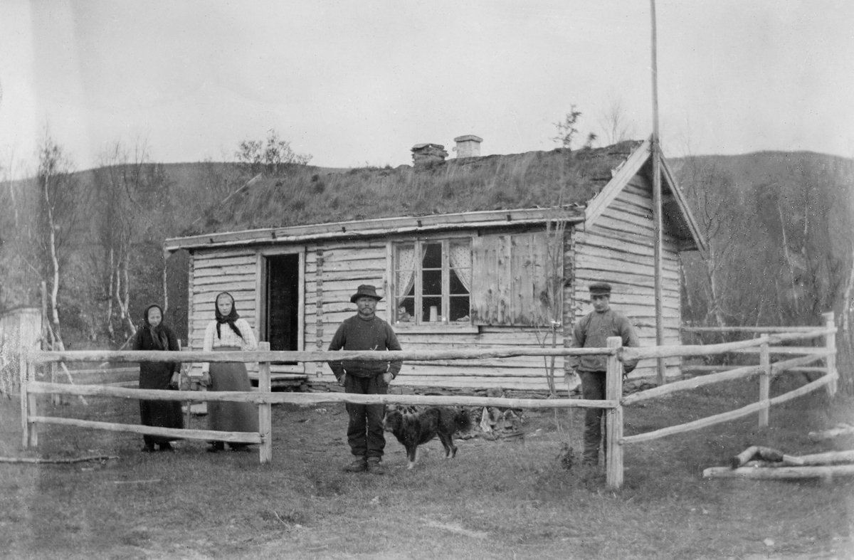 Skogstua ved Lulle i Skibotndalen. Fotografiet viser ei sinklaftet stue med torvtak og inngang på langsida.  Langveggen har også et trefags vindu med varelem.  Huset er inngjerdet av et rajegjerde.  To kvinner og to menn står mellom gjerdet og stua. 