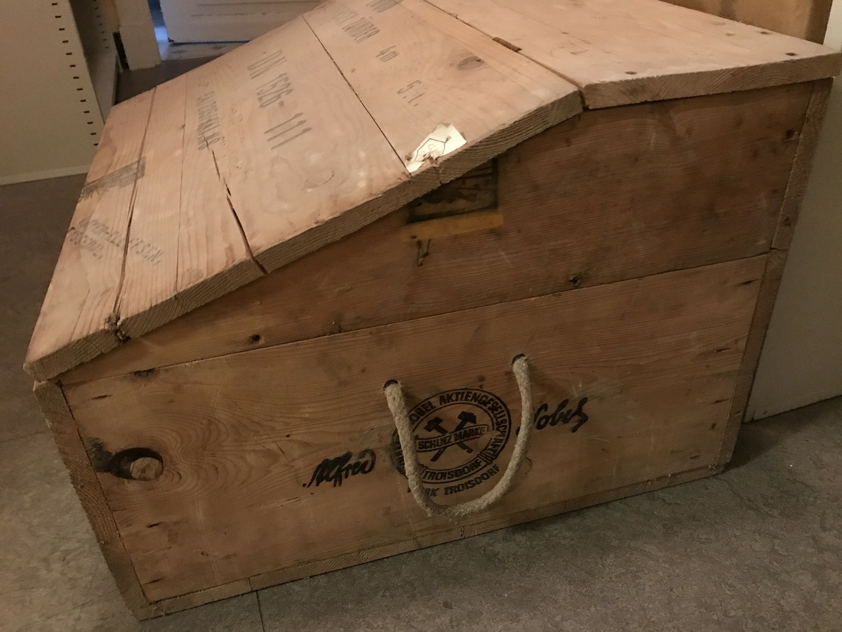 Trekasse med skrått, hengsla lok og hankar av fletta tau i to sider. Omarbeidd av to solide transportkassar for elektriske tennarar. Skrift og logo på lokk og sider. 