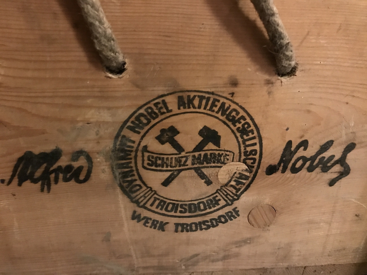 Trekasse med skrått, hengsla lok og hankar av fletta tau i to sider. Omarbeidd av to solide transportkassar for elektriske tennarar. Skrift og logo på lokk og sider. 