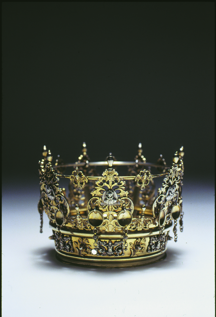 Brudkrona av förgyllt silver av Daniel Poppelman, Gävle 1770. Rikt dekorerad.
Ståndare i genombrutet arbete, 7st växtslingor med liten medaljong med renässanskvinnobild, bröstbild, i mitten. Upptill avlång topp.
På ståndarnas yttersidor hänger något nedom bildmedaljen två tunna, kupiga silverbrickor med repstav kring kanten och nertill avslutade med några länkar. Mellan ståndarnas påsatta ornament med vingad figur i genombrutet arbete; likaså vid ringens ovankant och på ringens sidor, här med rena bladornament emellan. På insidan av ringen ristat "58 1/2". Stämplar på yttrsidan av den nedre, smala ring, som håller ståndarna "G", tre kronor, "DPM" samt "M".