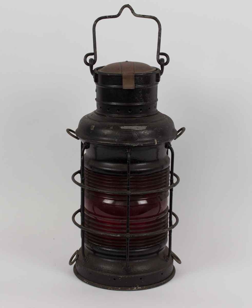 Rød lanterne, rundtlysende. Med hank på toppen og beskyttelsesgitter rundt glasset. Parafinbrenner mangler.