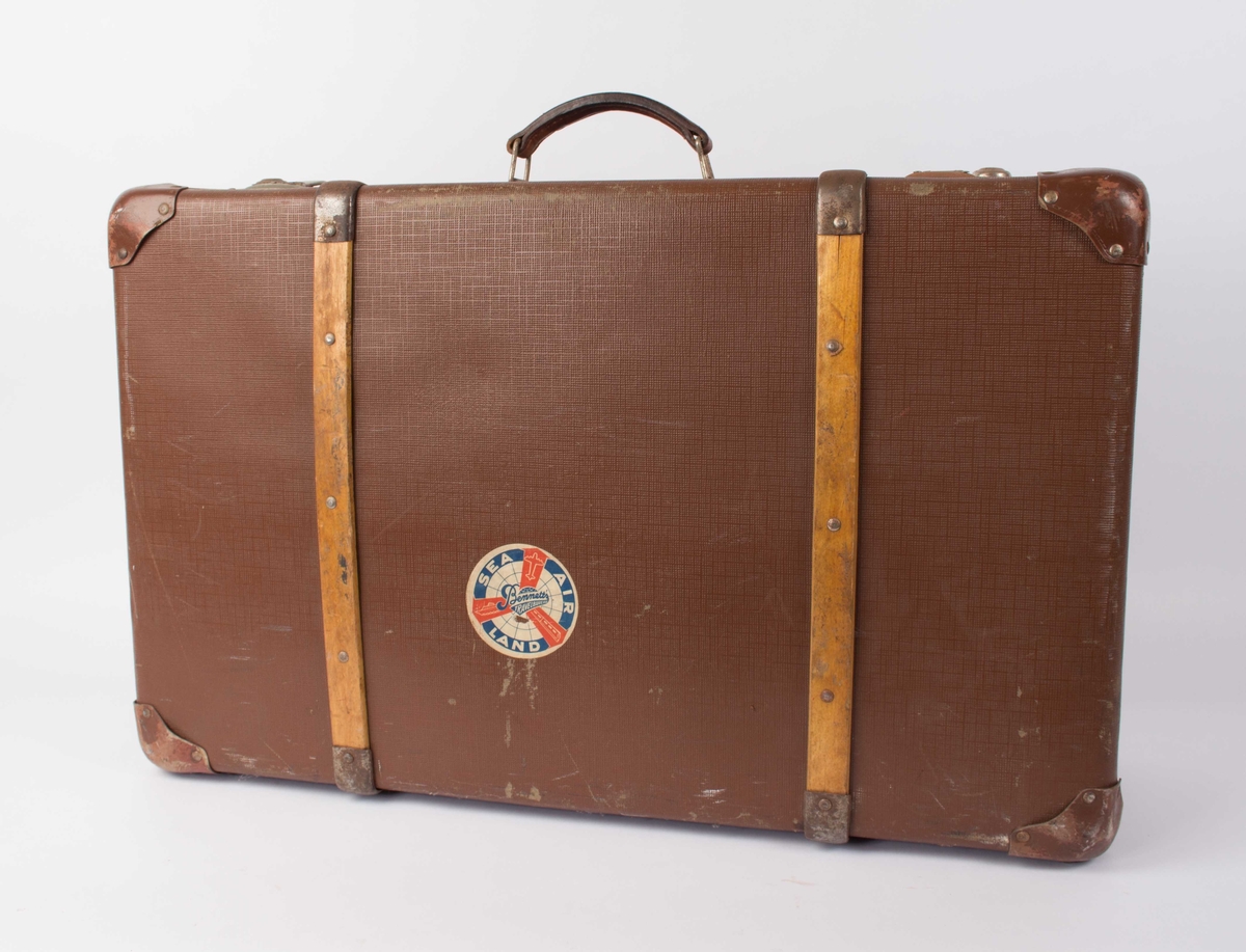 Brun vinylkoffert med trebeslag utvendig i bunn og topp. Hengslet lokk med med låsbeslag (nøkkel mangler)