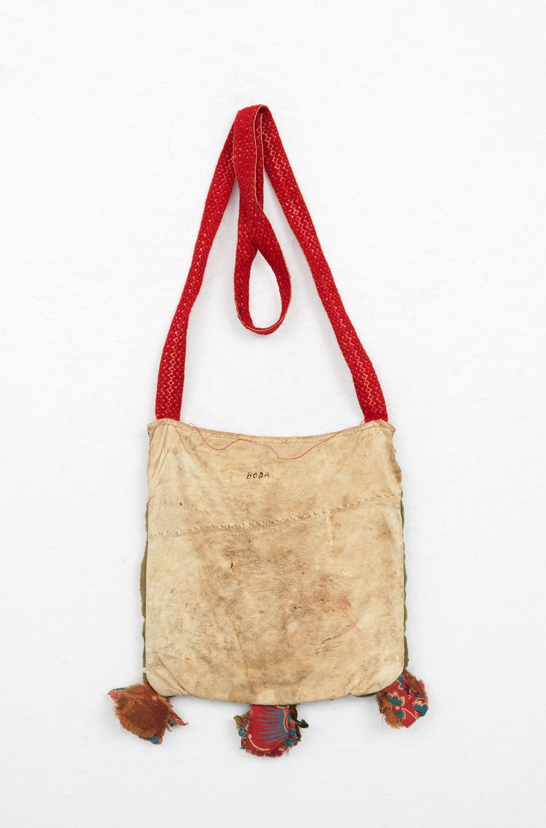 Kjolsäck, liduväska, till Boda sockendräkt för kvinna. Av vitt skinn med applikationer i grönt, gult och rött ylletyg, hjärtformer och blad. Broderier av silke och bomullsgarn i form av stjärnor och flätsömmar. På framstyckets överkant flätad dekor av smala skinn och tygremsor. Vid nederkanten små tofsar av tygbitar av olika kvaliteter. Passpoal av grönt ylletyg runt kanterna. Fickspegel av rödbottnad kattun. Baksida av vitt rakat skinn. Foder av kypertvävt linnetyg. Överkant på ficka och bakstycke kantat av vitt skinn. Vid överkantens hörn fastsytt ett mönstervävt band av rött ullgarn på vit botten. Märkning med initialer av rött ylletyg på framsidan: A H