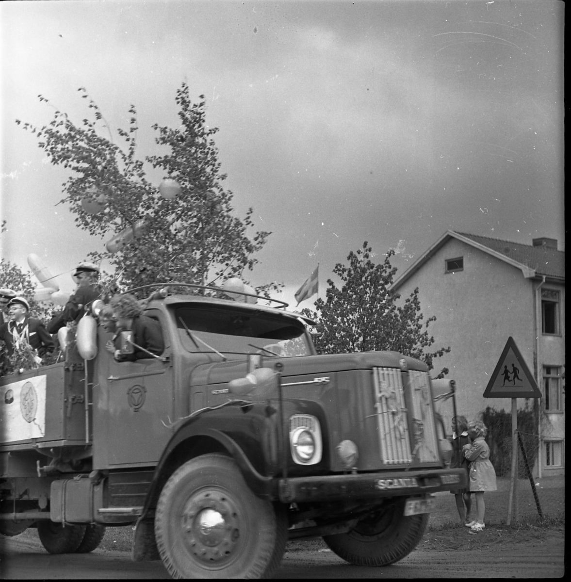 Realexamen 1961. Bilden föreställer ett lastbil, på flaket står flera studenter. De bär studentmössa och blombuketter runt sina halsar. Flaket är klätt med Björkris och ballonger. Två kvinnor sticker ut sina överkroppar genom fönstret på passagerarsidan på lastbilen.