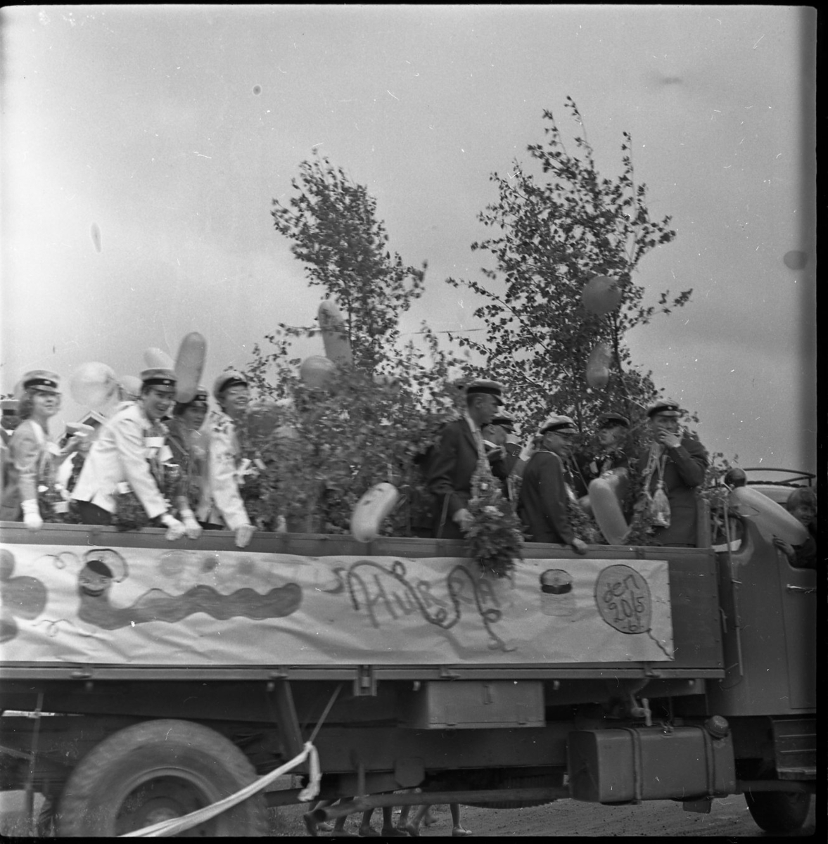 Realexamen 1961. Bilden föreställer ett lastbilsflak, på flaket står flera studenter. De bär studentmössa och blombuketter runt sina halsar. Flaket är klätt med Björkris och ballonger.