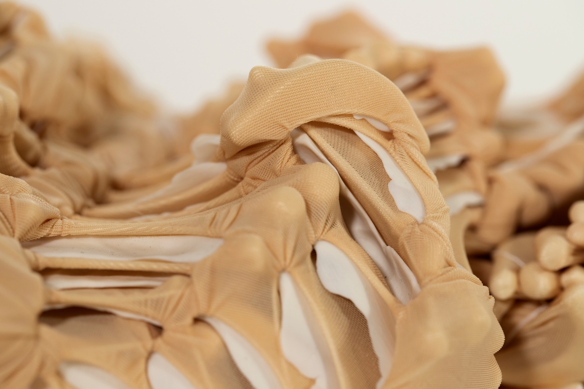 Objekt/ skulptur med organisk form. Beige nylon er knyttet og sydd rundt porselensskiver og foldet. Formen kan minne om et kadaver.