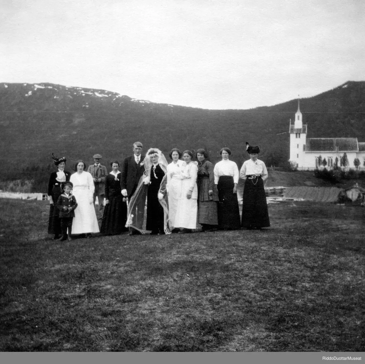 Náittosbárra ja guossit girkugieddis, girku duogábealde.
Brudepar med gjester, kirka i bakgrunnen