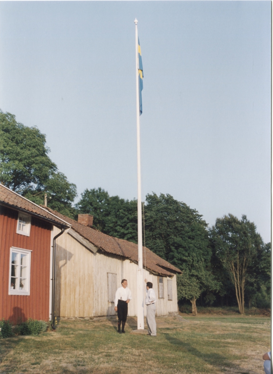 Staffan Bjerrhede och Berny Gustafsson har hissat flaggan inför hembygdsgårdens midsommarfirande 1993. Från vänster ses Hembygdsgården Långåker 1:3 och Långåker 1:2 "Davids", "Derras".
Relaterade motiv: A03389 - A03408.