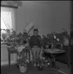 Realskolebilder Nyarp 1961. En kvinna sitter på en stol omgiven av blommor. Hon bär studentmössa och ser glad ut.