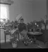 Realskolebilder Nyarp 1961. En kvinna sitter på en stol omgiven av blommor. Hon bär studentmössa och ser glad ut.