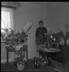 Realskolebilder Nyarp 1961. En kvinna står omgiven av krukväxter och blombuketter. Hon bär studentmössa och ser glad ut.