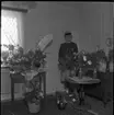 Realskolebilder Nyarp 1961. En kvinna står omgiven av krukväxter och blombuketter. Hon bär studentmössa.