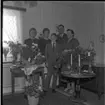 Realskolebilder Nyarp 1961. En familj står och poserar för en gruppbild. Omkring dem står krukväxter och blomuketter utställda. På ett bord står två ljus.