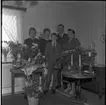 Realskolebilder Nyarp 1961. En familj står och poserar för en gruppbild. Omkring dem står krukväxter och blomuketter utställda. På ett bord står två ljus.