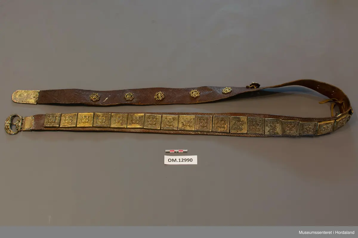 Stølabelte og fangreim i eitt. Eit langt lærstykke der det i eine halvdelen er montert 19 firkanta stølar, og i den andre enden 6 runde, små, blomsterliknande borer. Dei ulike delane har festeringar som går gjennom hol i leret, og vert festa på baksida ved at ei snor av bomull vert trekt gjennom ringane. Endestykke nedst på fangreima. På baksida er nokre svake merker: Nokre X-ar og ein L. Beltet har mykje restar av pussemiddel.