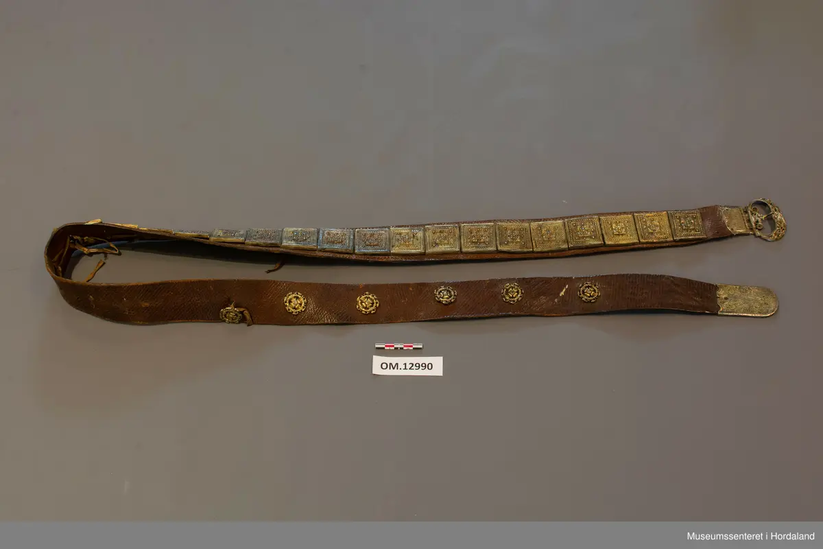 Stølabelte og fangreim i eitt. Eit langt lærstykke der det i eine halvdelen er montert 19 firkanta stølar, og i den andre enden 6 runde, små, blomsterliknande borer. Dei ulike delane har festeringar som går gjennom hol i leret, og vert festa på baksida ved at ei snor av bomull vert trekt gjennom ringane. Endestykke nedst på fangreima. På baksida er nokre svake merker: Nokre X-ar og ein L. Beltet har mykje restar av pussemiddel.