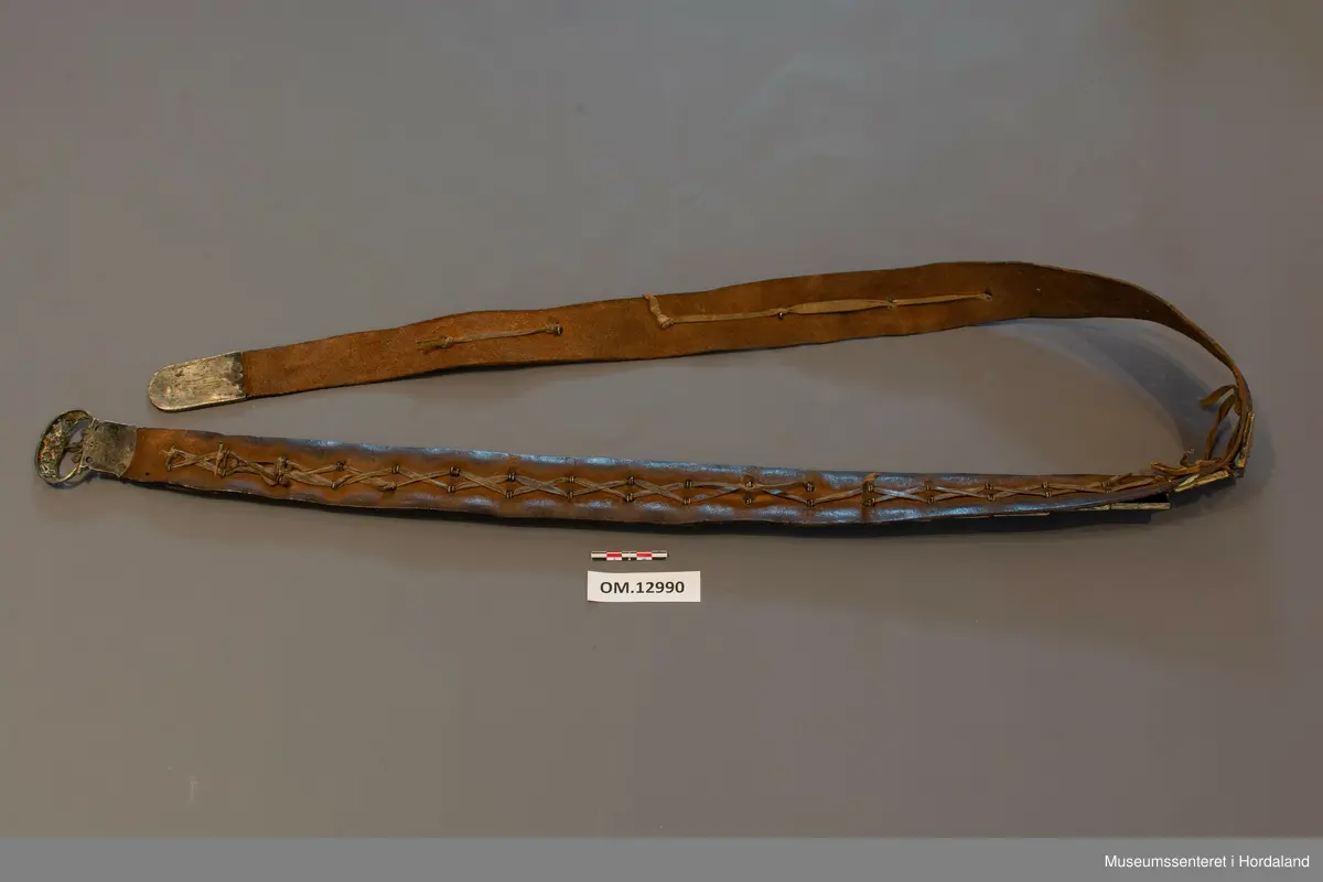 Stølabelte og fangreim i eitt. Eit langt lærstykke der det i eine halvdelen er montert 19 firkanta stølar, og i den andre enden 6 runde, små, blomsterliknande borer. Dei ulike delane har festeringar som går gjennom hol i leret, og vert festa på baksida ved at ei snor av bomull vert trekt gjennom ringane. Endestykke nedst på fangreima. På baksida er nokre svake merker: Nokre X-ar og ein L. Beltet har mykje restar av pussemiddel.