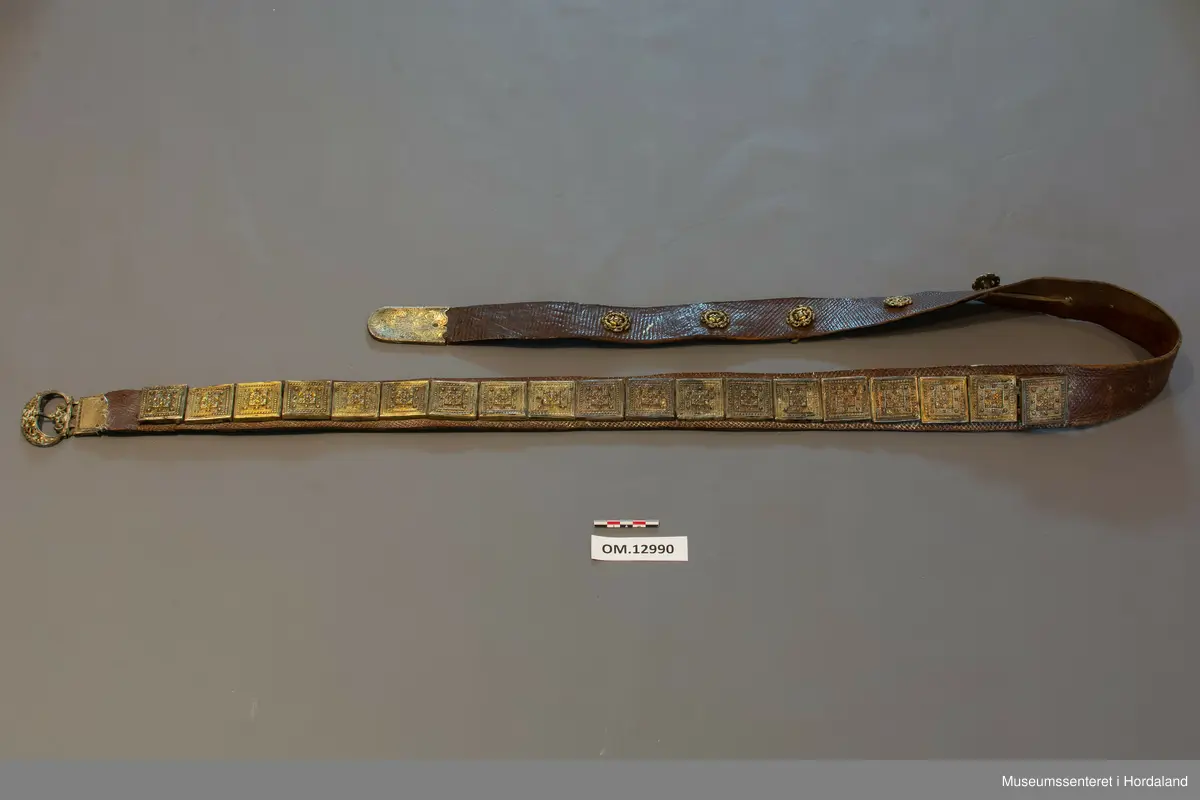 Stølabelte og fangreim i eitt. Eit langt lærstykke der det i eine halvdelen er montert 19 firkanta stølar, og i den andre enden 6 runde, små, blomsterliknande borer. Dei ulike delane har festeringar som går gjennom hol i leret, og vert festa på baksida ved at ei snor av bomull vert trekt gjennom ringane. Endestykke nedst på fangreima. På baksida er nokre svake merker: Nokre X-ar og ein L. Beltet har mykje restar av pussemiddel.