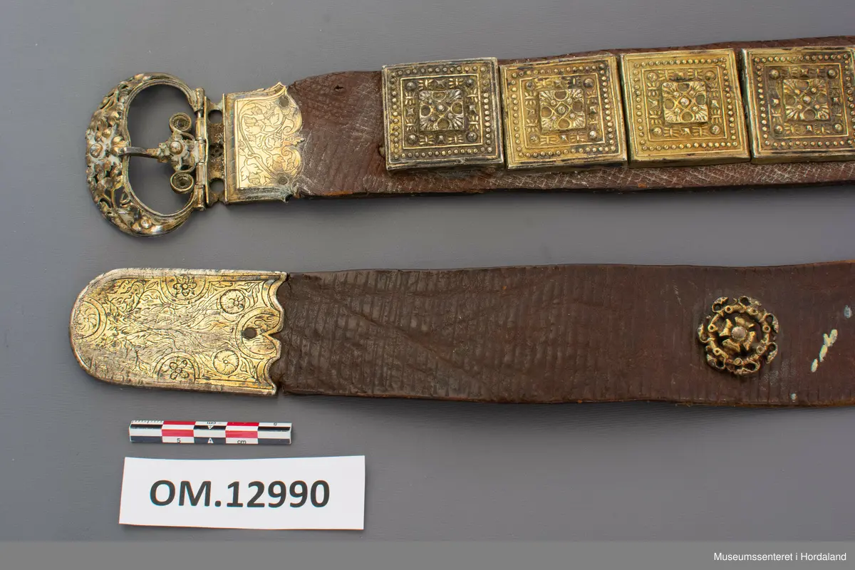 Stølabelte og fangreim i eitt. Eit langt lærstykke der det i eine halvdelen er montert 19 firkanta stølar, og i den andre enden 6 runde, små, blomsterliknande borer. Dei ulike delane har festeringar som går gjennom hol i leret, og vert festa på baksida ved at ei snor av bomull vert trekt gjennom ringane. Endestykke nedst på fangreima. På baksida er nokre svake merker: Nokre X-ar og ein L. Beltet har mykje restar av pussemiddel.