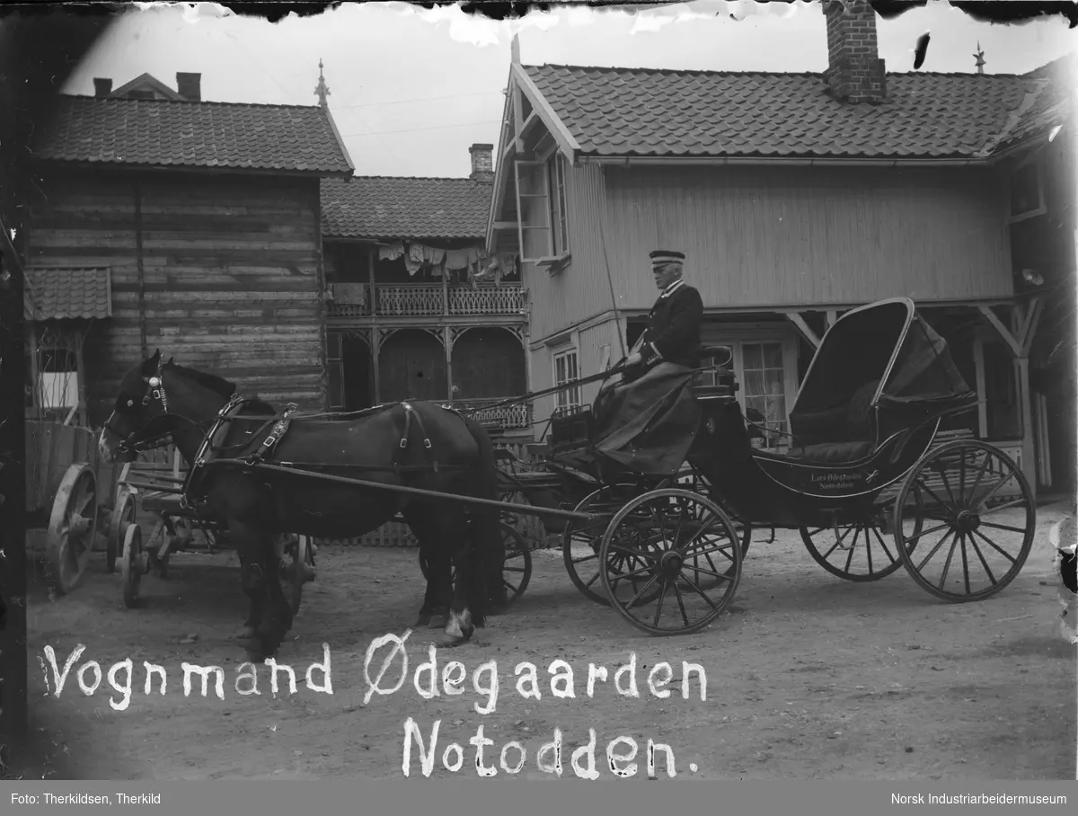 To hester med seletøy spent for vogn, kuskesete foran på vogna. Vognmann Lars Ødegaard sitter på kuskesete med klede over beinene. Bygninger i bakgrunnen. Svalgang der klær henger til tørk.