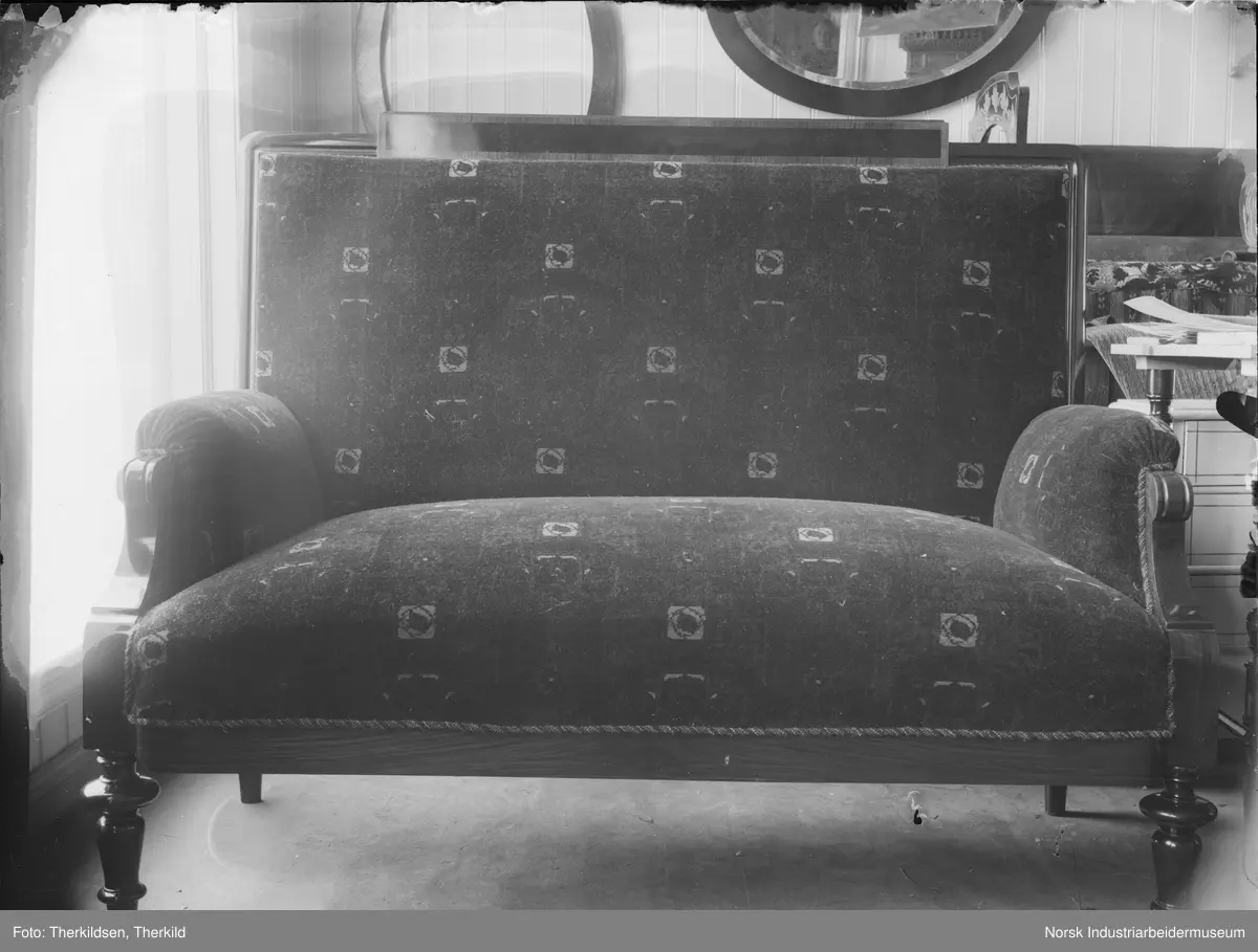 Sofa med treben og tredetaljer på armlene. Speil på veggen i bakgrunnen, kvinne sees i speilet.