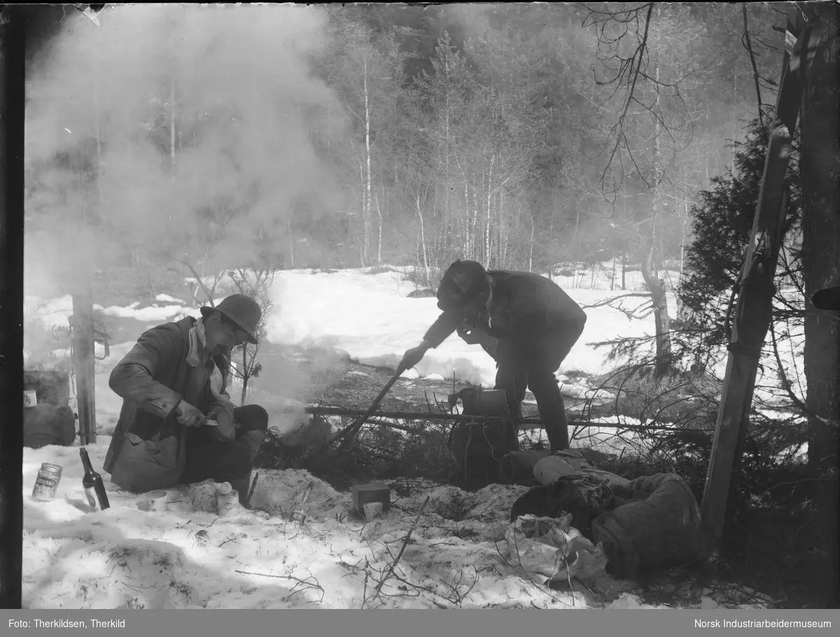 To menn på skitur raster i skogen med bål. Boksmat og flasker står i snøen rundt. Sekker med utstyr står inntil skiene. Begge mennene med pipe i munnen.