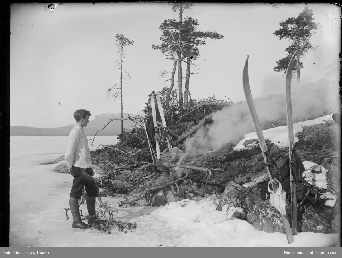Mann stående og ser på bål. Et par ski på hver side av bålet. Mannen står på det islagte vannet Follsjø, ved Leirodden.