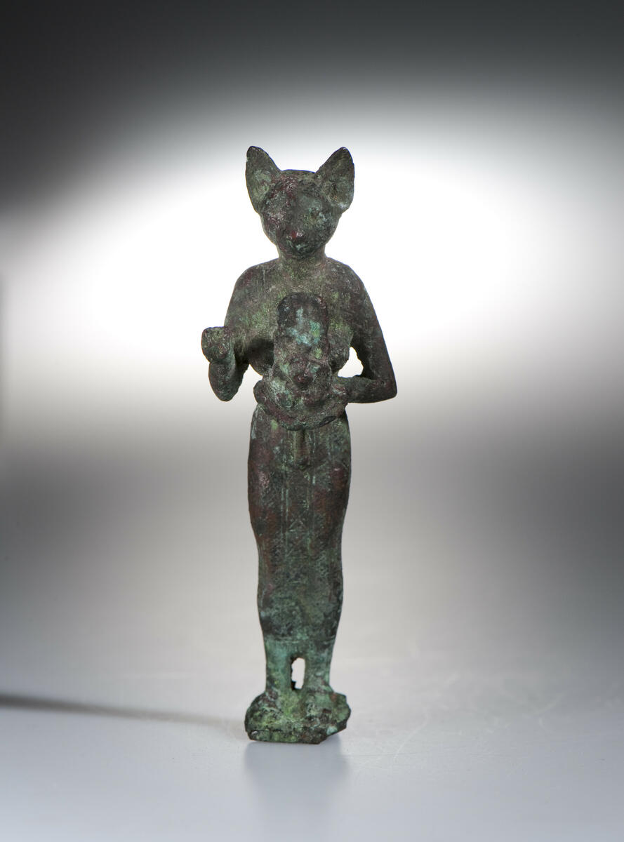 Figurin av brons, Egypten- Egyptisk gudabild föreställande Gudinnan Bastet, symboliserad som en katt. Daterad till Setiden, 664-525 f.kr. Längd: 9 cm, Höjd: 2 cm, Bredd: 2,7 cm.  Gudinnan Bastet har här avbildats med ett katthuvud på en kvinnokropp. I sin vänstra hand håller hon en ”aegis”, en skyddssköld med ett katt- eller Sekhmetansikte (krigets och solens gudinna med lejonhuvud på en kvinnorkropp) med en bred krage under. I sin högra hand har hon troligtvis hållit ett ”sistrum”, ett musikinstrument med små metallplattor vilka rasslade när man skakade instrumentet. Sistrumet är dock bortslaget. Statyetten har antagligen varit fastsatt på en träsockel.; ; Under Sentiden tillverkades små bronsfigurer i stora mängder och antagligen är denna statyett ett exempel på dessa. De skänktes därefter som votivgåvor till olika tempel. Tillverkningstid: 664-525 k Kr.