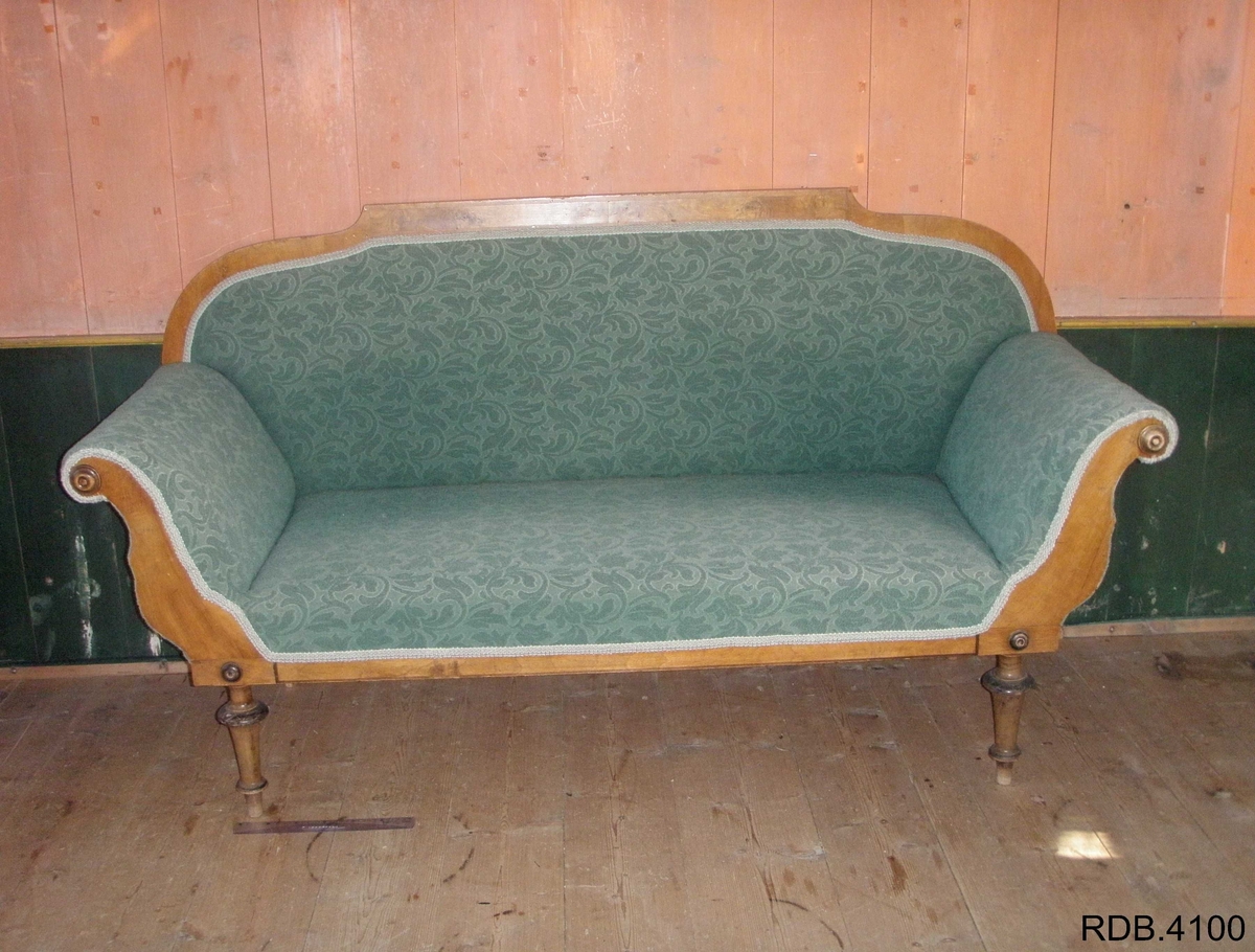 En 3-seters sofa. Rygg og sete av stoff, føtter og kanter av tre. Fire, runde føtter. Stoffet er i et grønt blomstermønster. Et armlene på hver side, som skrår utover. To runde detaljer på hver av armlenene. 