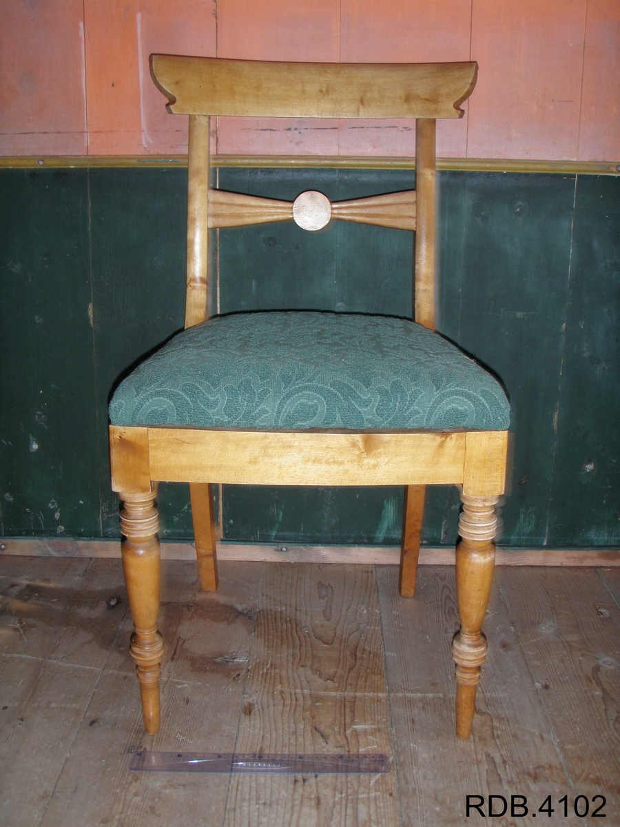 Stol i tre, lakkert. Fire føtter; framføttene er runde/dreiet, mens bakføttene er firkantede. Alle føtter er bredest øverst, og smalest nederst. Bakføttene fortsetter oppover, og er en del av stolryggen. Stolryggen har en støtte øverst, og en støtte midt på. Støtten øverst er firkantet. Støtten midt på har en runding midt på, og mot sidene går det to firkanter. 
Stolputa er løs, men sitter godt på fordi den er festet til noen spor på undersiden. Den er bredest frampå, og smalest bakerst. Stolputa er av tre, og trekt med et fôret, grønt stoff. 