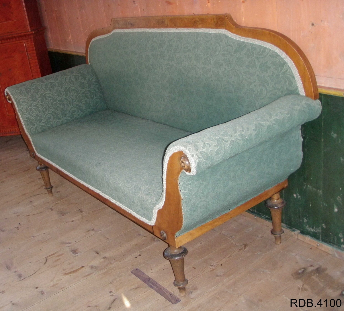 En 3-seters sofa. Rygg og sete av stoff, føtter og kanter av tre. Fire, runde føtter. Stoffet er i et grønt blomstermønster. Et armlene på hver side, som skrår utover. To runde detaljer på hver av armlenene. 