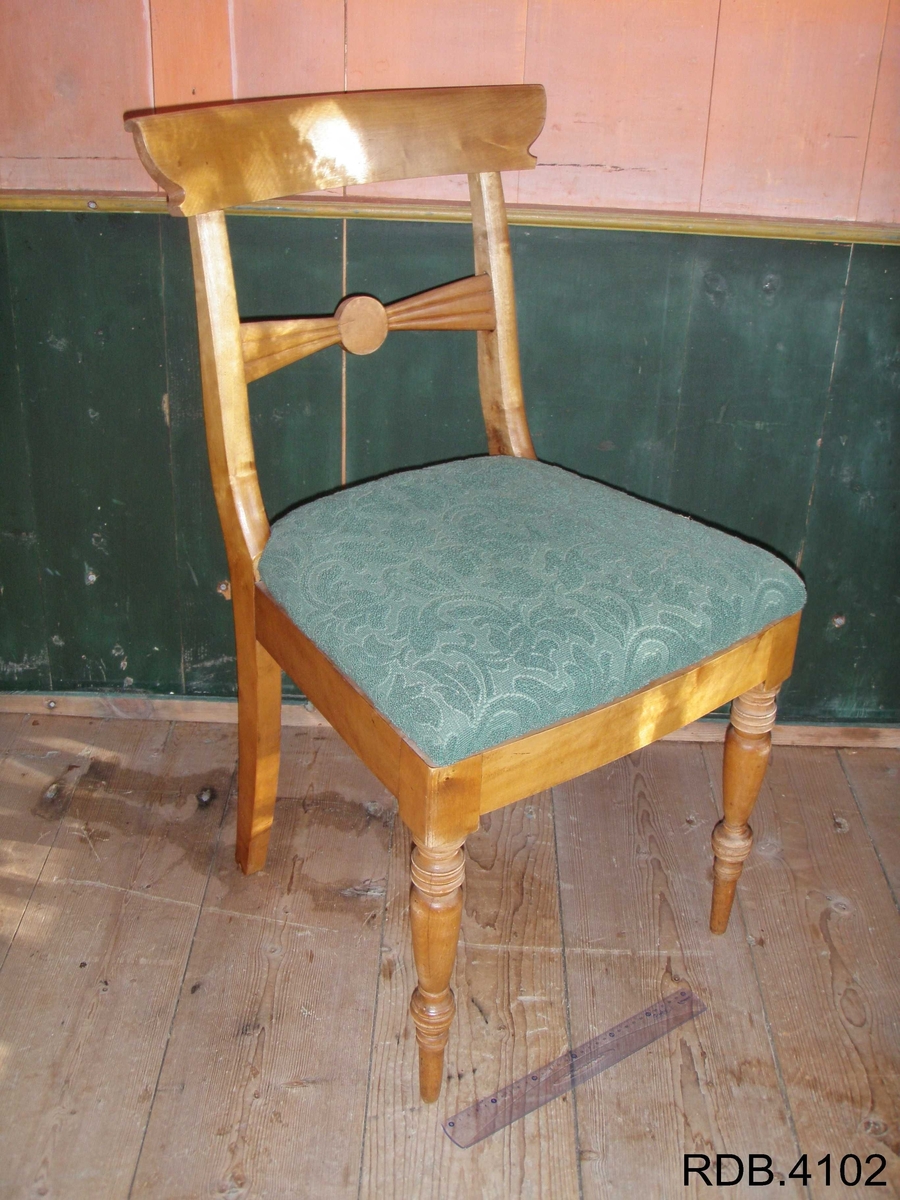 Stol i tre, lakkert. Fire føtter; framføttene er runde/dreiet, mens bakføttene er firkantede. Alle føtter er bredest øverst, og smalest nederst. Bakføttene fortsetter oppover, og er en del av stolryggen. Stolryggen har en støtte øverst, og en støtte midt på. Støtten øverst er firkantet. Støtten midt på har en runding midt på, og mot sidene går det to firkanter. 
Stolputa er løs, men sitter godt på fordi den er festet til noen spor på undersiden. Den er bredest frampå, og smalest bakerst. Stolputa er av tre, og trekt med et fôret, grønt stoff. 