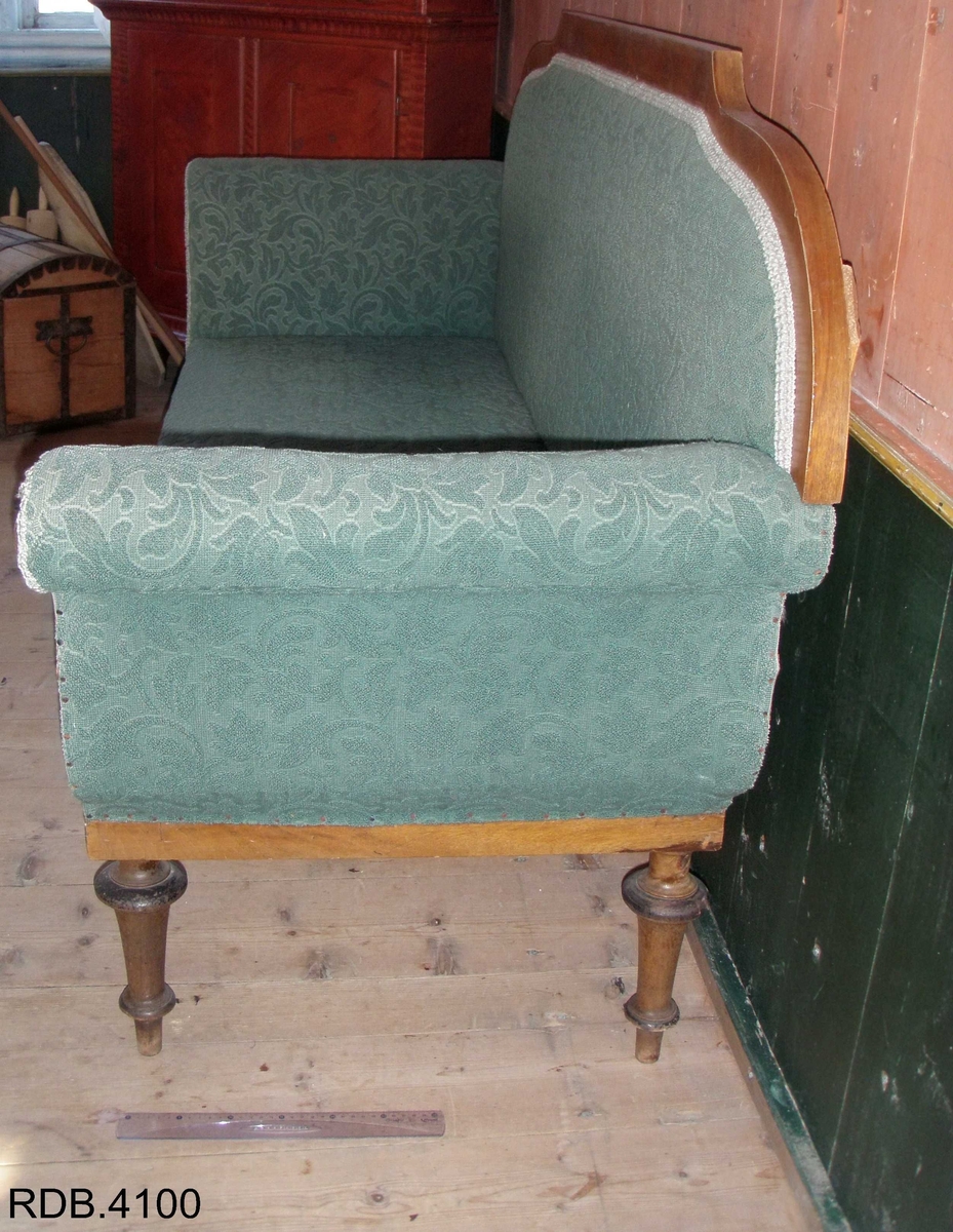 En 3-seters sofa. Rygg og sete av stoff, føtter og kanter av tre. Fire, runde føtter. Stoffet er i et grønt blomstermønster. Et armlene på hver side, som skrår utover. To runde detaljer på hver av armlenene. 