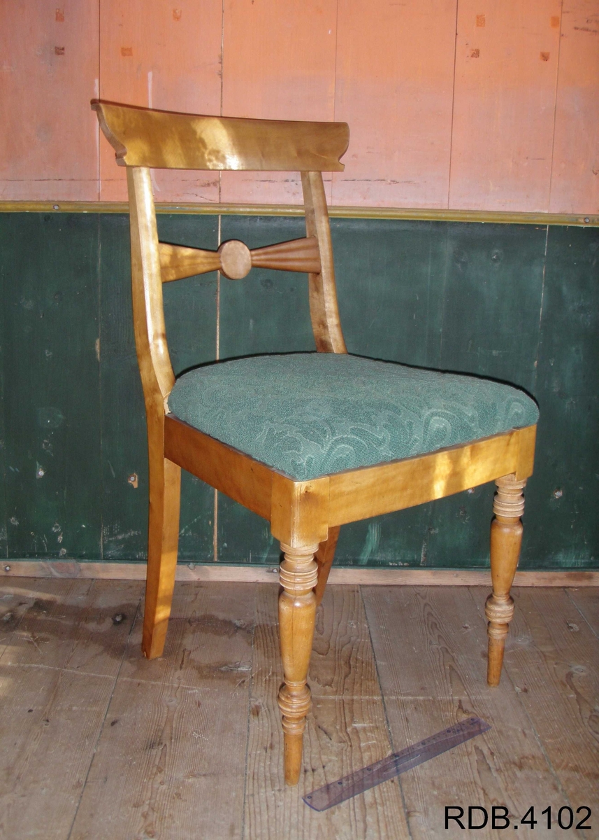 Stol i tre, lakkert. Fire føtter; framføttene er runde/dreiet, mens bakføttene er firkantede. Alle føtter er bredest øverst, og smalest nederst. Bakføttene fortsetter oppover, og er en del av stolryggen. Stolryggen har en støtte øverst, og en støtte midt på. Støtten øverst er firkantet. Støtten midt på har en runding midt på, og mot sidene går det to firkanter. 
Stolputa er løs, men sitter godt på fordi den er festet til noen spor på undersiden. Den er bredest frampå, og smalest bakerst. Stolputa er av tre, og trekt med et fôret, grønt stoff. 